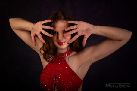 Frau mit lockigem Haar in rotem Kleid, die spielerisch ihre Hände vor dem Gesicht hält.