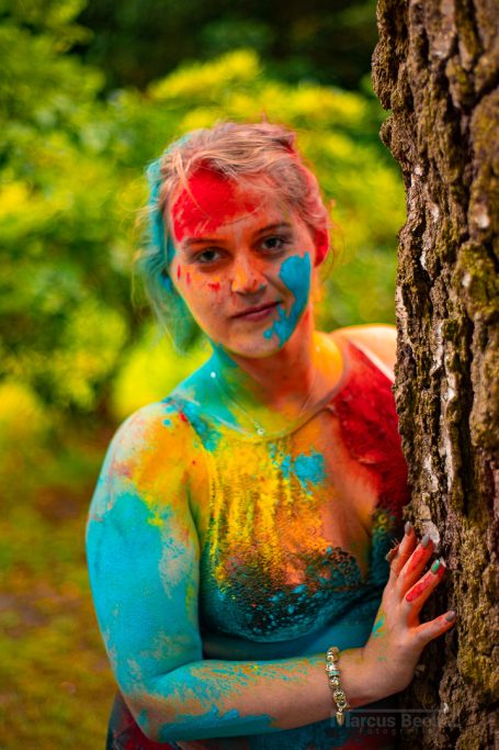 Frau mit buntem Körperfarben in einem Wald, neben einem Baum stehend.