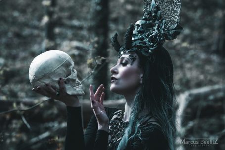 Frau mit grünem Haar hält einen menschlichen Schädel in einem mystischen Wald.