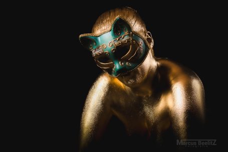 Eine bemalte Person mit einer Katzenmaske und goldener Körperfarbe auf dunklem Hintergrund.
