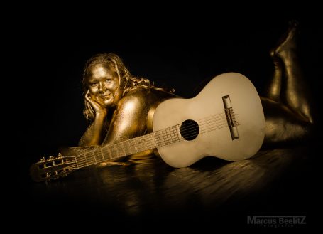 Frau in Gold mit Gitarre, entspannt liegend auf einem dunklen Hintergrund.