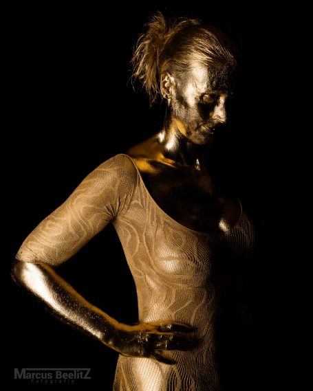 Eine person in einem eng anliegenden Oberteil, mit goldener Körperbemalung und nachdenklichem Ausdruck.
