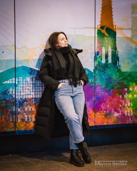 Frau in schwarz-grauem Outfit lehnt an bunter Wand mit städtischem Hintergrund.
