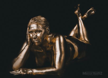 Eine Person mit goldfarbener Körperbemalung liegt auf dem Bauch und stützt den Kopf auf die Hand.