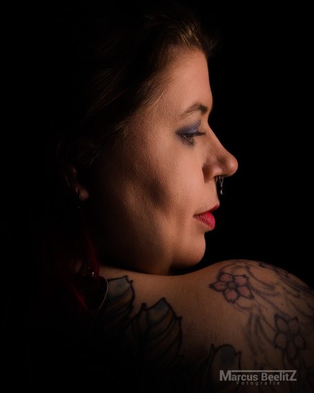 Profil einer Frau mit roten Lippen und Tattoos auf dem Schultern, vor dunklem Hintergrund.