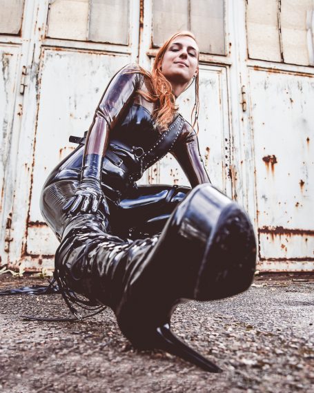 Frau in schwarzem, knallengen Latex-Outfit und hohen Stiefeln, posiert vor einer grauen Wand.