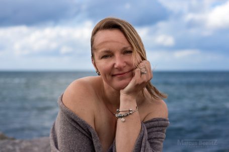Eine Frau mit schulterlangen blonden Haaren lächelt am Meer, im Hintergrund ein bewölkter Himmel.