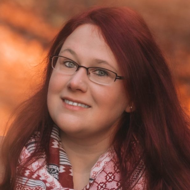 Frau mit langen, roten Haaren und Brille, lächelt vor einem unscharfen, herbstlichen Hintergrund.
