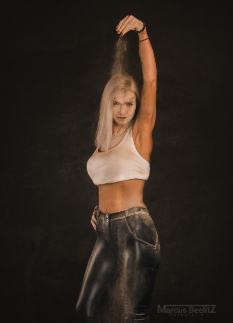 Frau mit blonden Haaren, die Sand über ihre Schulter streut, in einem körperbetonten Outfit.