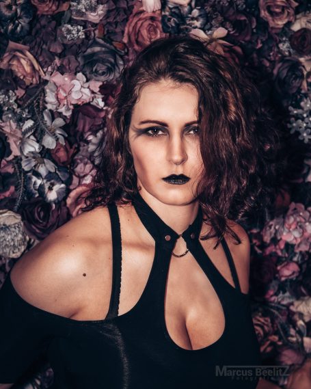 Eine Frau mit düsterem Make-up steht vor einer Wand aus Blumen.