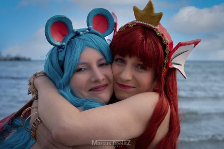 Zwei freundlich lächelnde Frauen in fantasievollen Kostümen umarmen sich am Strand.