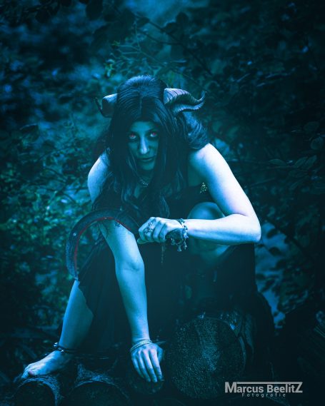 Frau mit langen Haaren in einem dunklen, mystischen Wald, in nachdenklicher Pose.