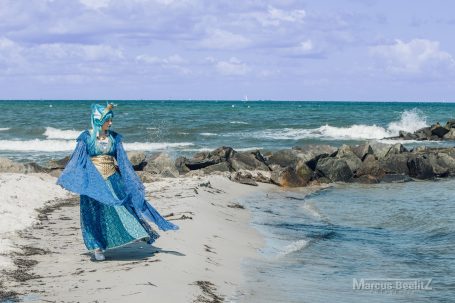 Person in einem blauen Kostüm steht am Strand mit Wellen im Hintergrund.