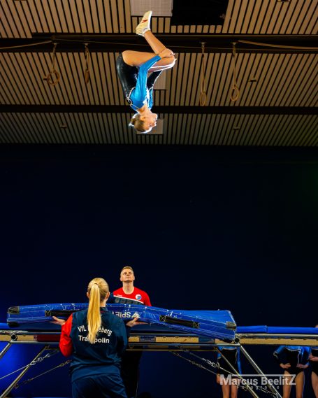 Akrobat springt hoch über ein Trampolin, während Trainer und Zuschauer beobachten.