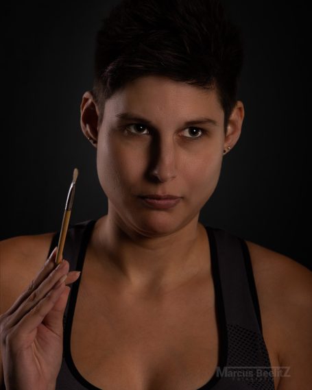 Junge Frau mit kurzem Haar hält einen Pinsel in der Hand, schaut ernst in die Kamera.