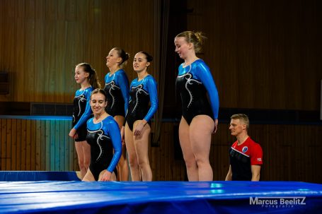 Fünf Turnerinnen in Gymnastikleotards stehen auf einem Sprungtisch, ein Trainer daneben.