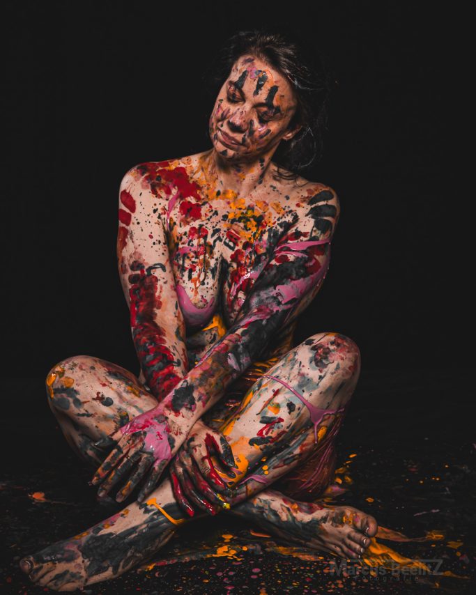 Eine sitzende Person, bemalt mit bunten Farben, auf einem dunklen Hintergrund.