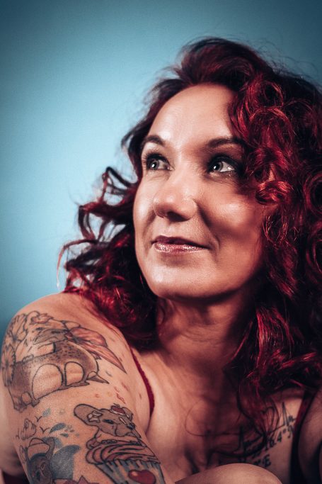 Frau mit lockigem rotem Haar, Tattoos auf dem Oberkörper, blickt nachdenklich zur Seite.