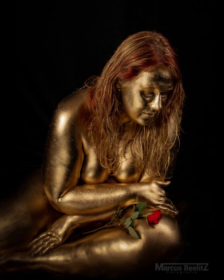Eine goldene, sitzende Frau hält eine rote Rose in einer dunklen Umgebung.