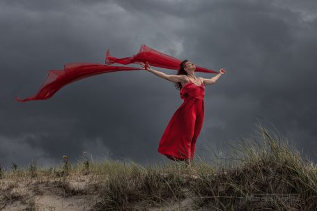 Frau in einem roten Kleid mit wehenden Tüchern vor einem düsteren Himmel.