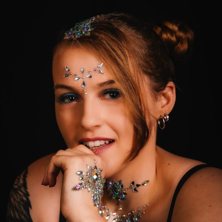 Frau mit dekorativen Glitzersteinen im Haar und auf der Haut, tätowierter Arm sichtbar.