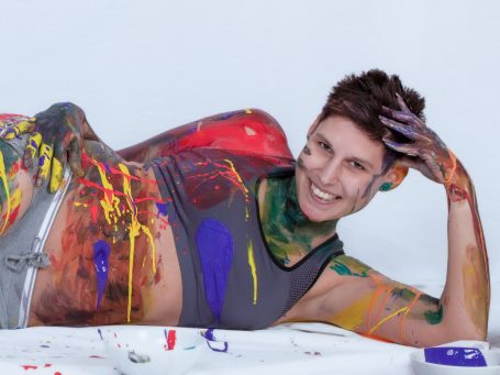 Eine lachende Frau mit bemalter Haut liegt entspannt auf einem weißen Hintergrund.